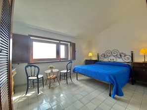A bedroom with a bed, two chairs, a small table with a plant, a window with blinds, and a nightstand with a lamp.