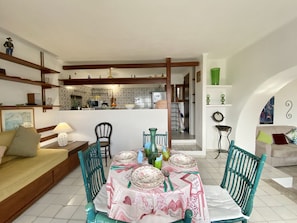 Un coin repas avec une table dressée pour quatre personnes, entourée de chaises de couleur turquoise. On aperçoit une cuisine à l’arrière-plan, avec un mur recouvert de carreaux.