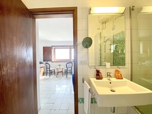 A bathroom with a sink, mirror, and shower. There is a window with blinds and a door leading to a dining area with chairs and a table.