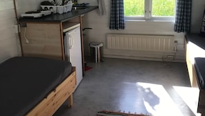Una habitación compacta con una cama, un escritorio pequeño con utensilios de cocina, un radiador y una ventana con cortinas.