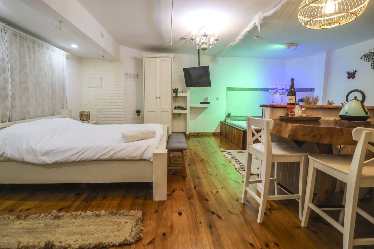 A bedroom with a bed, bedside table, and a small bench. There is a TV mounted on the wall, a wooden floor, and a dining area with a table and chairs.