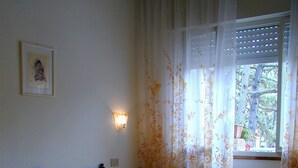 A bedroom with a bed, a nightstand, a wall-mounted light, and a window with curtains. - Villa Anny apartment 1 (Lignano Sabbiadoro)