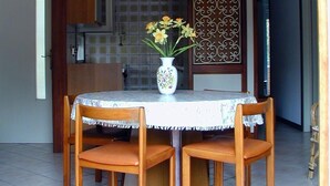 A dining area with a round table, four wooden chairs, a vase with flowers, and a decorative wall panel. - Villa Anny apartment 1 (Lignano Sabbiadoro)