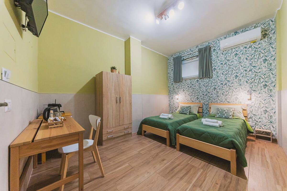 A room with two beds, a desk, a chair, a wardrobe, and an air conditioning unit.