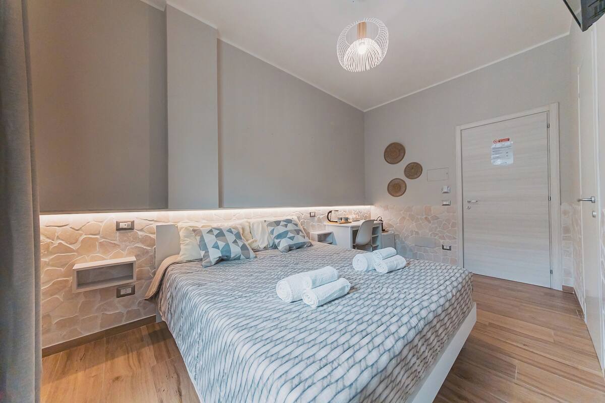 A bedroom with a stone accent wall, a bed with a patterned bedspread, two bedside tables, a desk, and a chair.