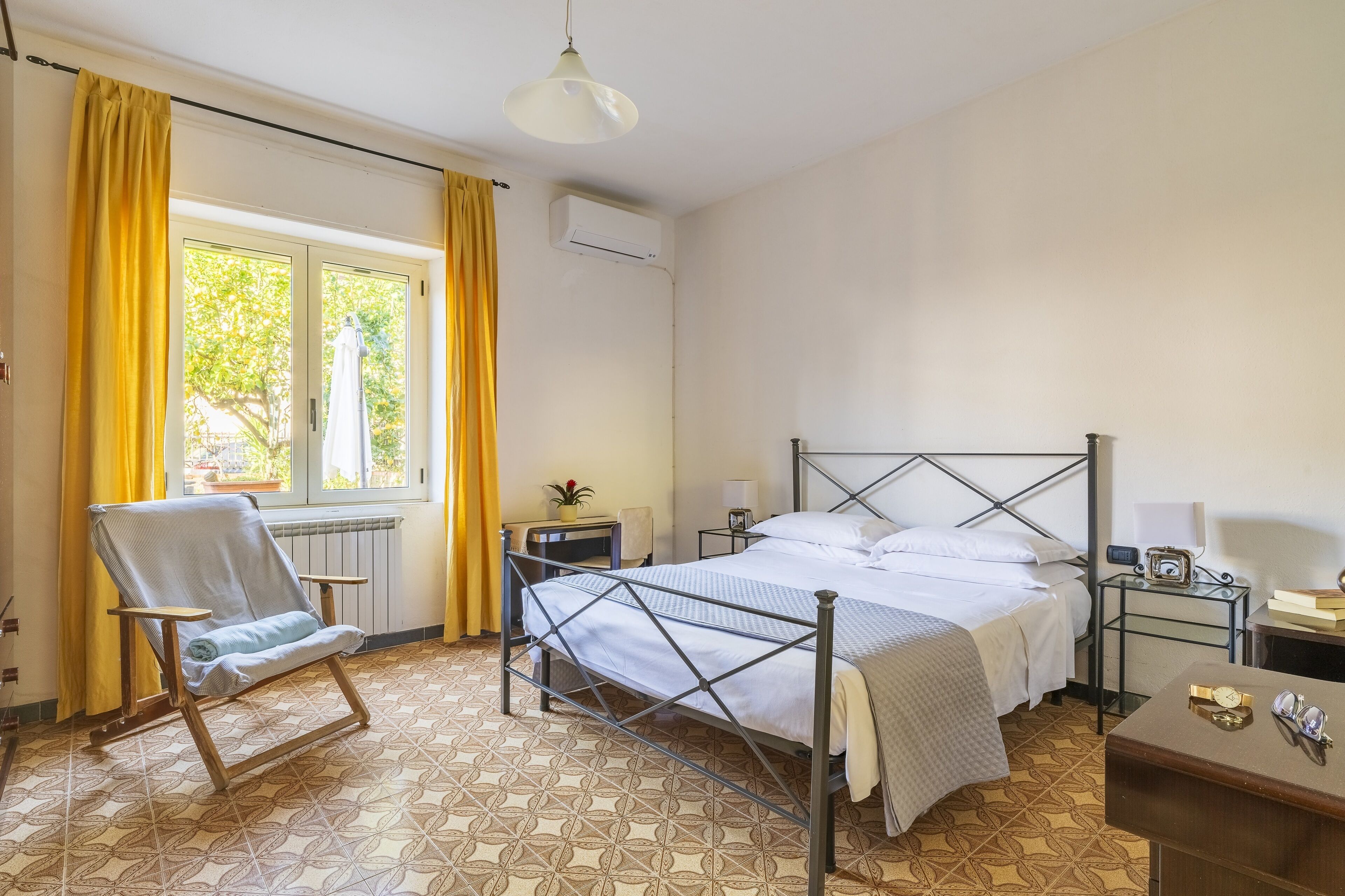 A bedroom with a metal-framed bed, a chair, a bedside table, and a window with curtains.
