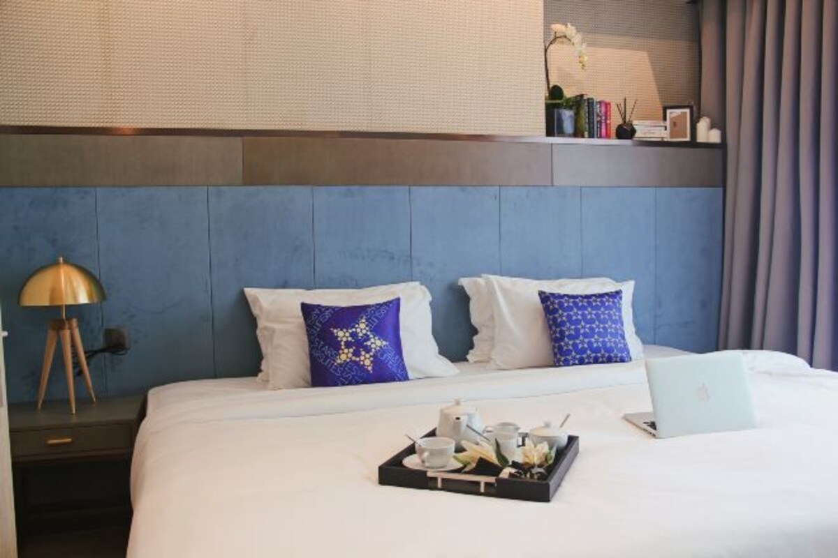 Habitación de hotel con cama, una laptop, una bandeja con tazas y tetera, y una mesita de noche con lámpara.