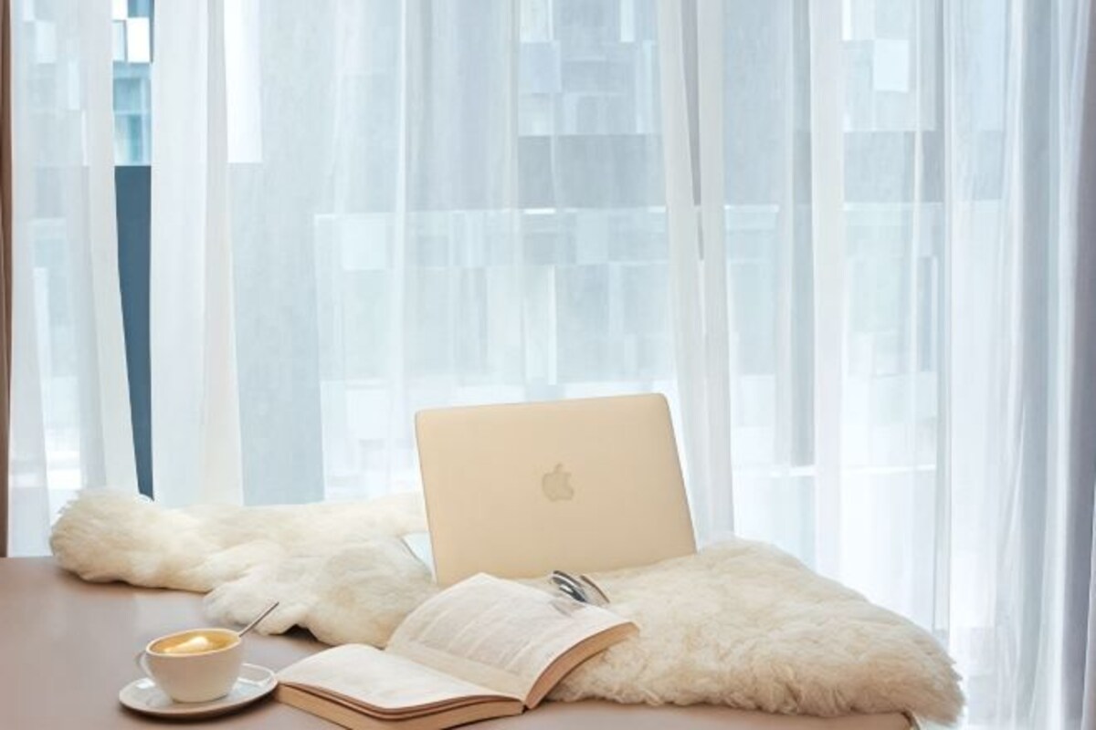 Una laptop sobre un cojín mullido, un libro abierto y una taza de café en una mesa junto a una ventana con cortinas translúcidas.