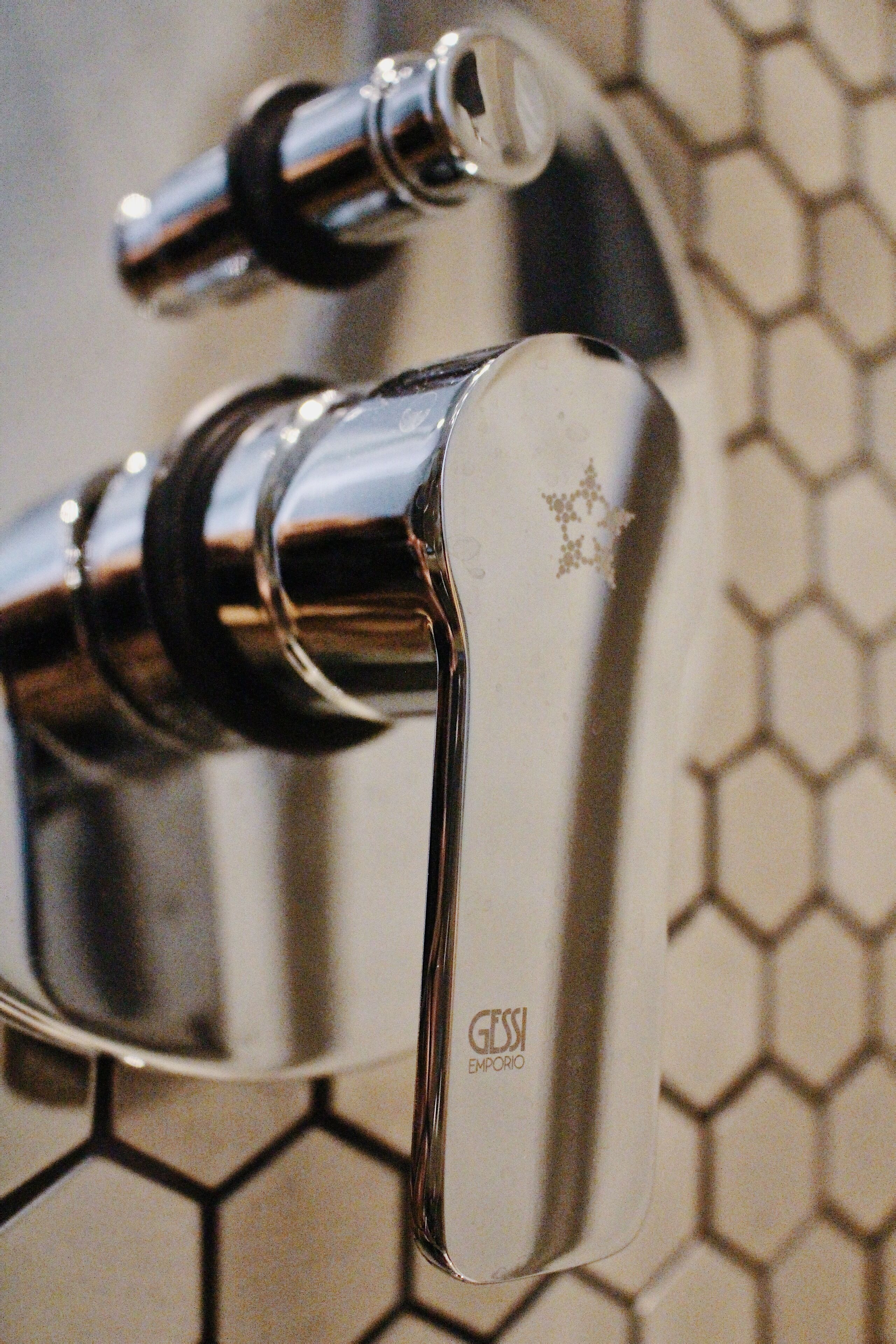 A chrome faucet with a visible brand logo, set against a tiled wall with a honeycomb pattern.