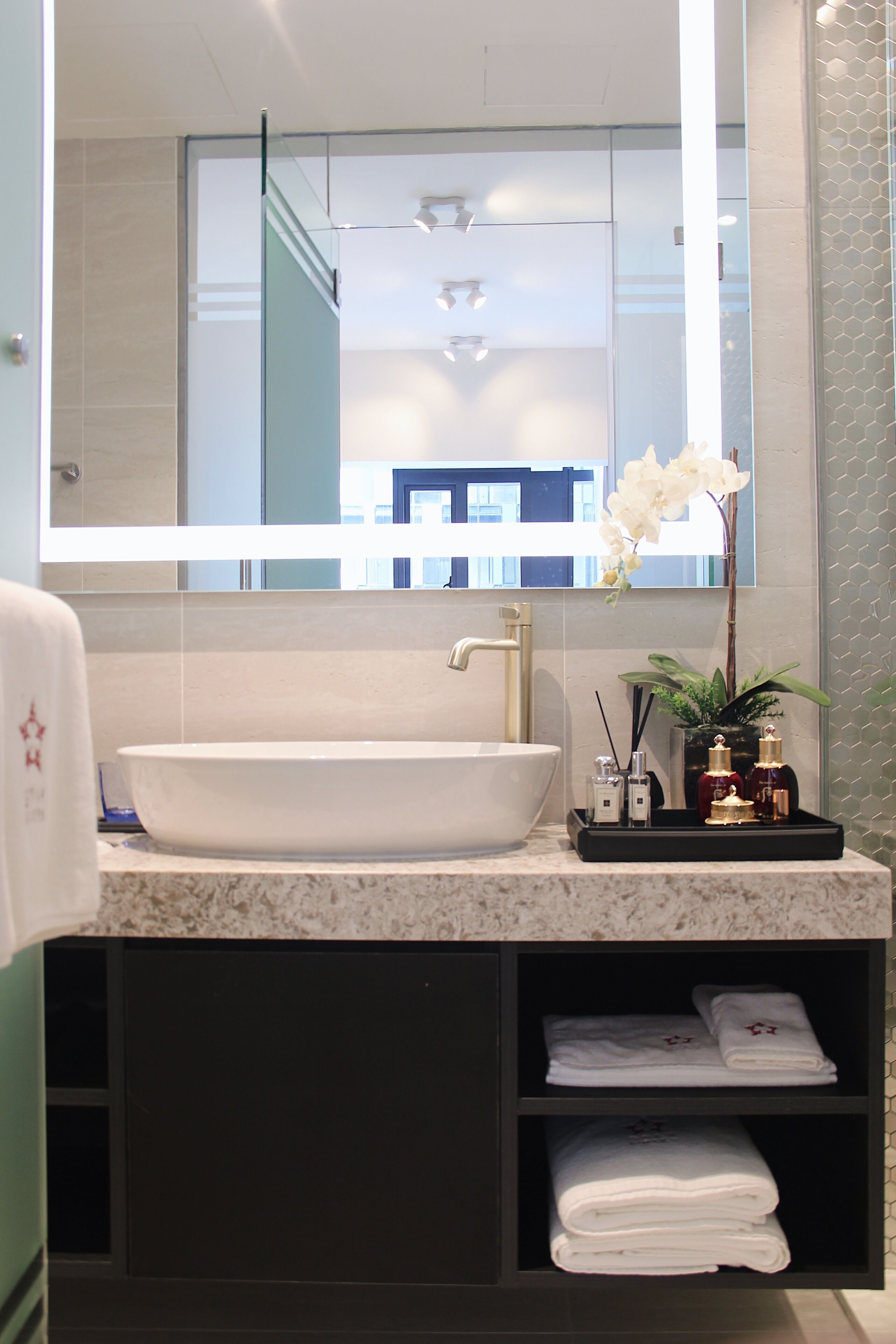 A bathroom with a large white sink, a mirror, and a potted orchid. There are two shelves below the sink, one holding folded towels and the other containing toiletries.