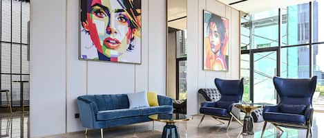 A modern lobby with a blue sofa, a round coffee table, and two armchairs. There are large windows and artwork on the walls.