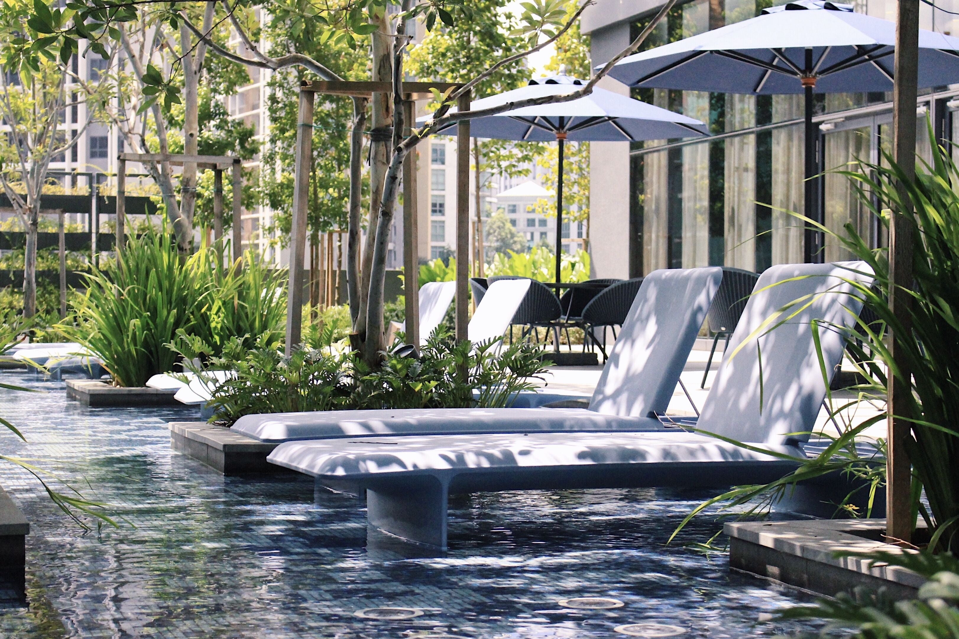 A pool area with lounge chairs and umbrellas, surrounded by greenery and modern buildings in the background.