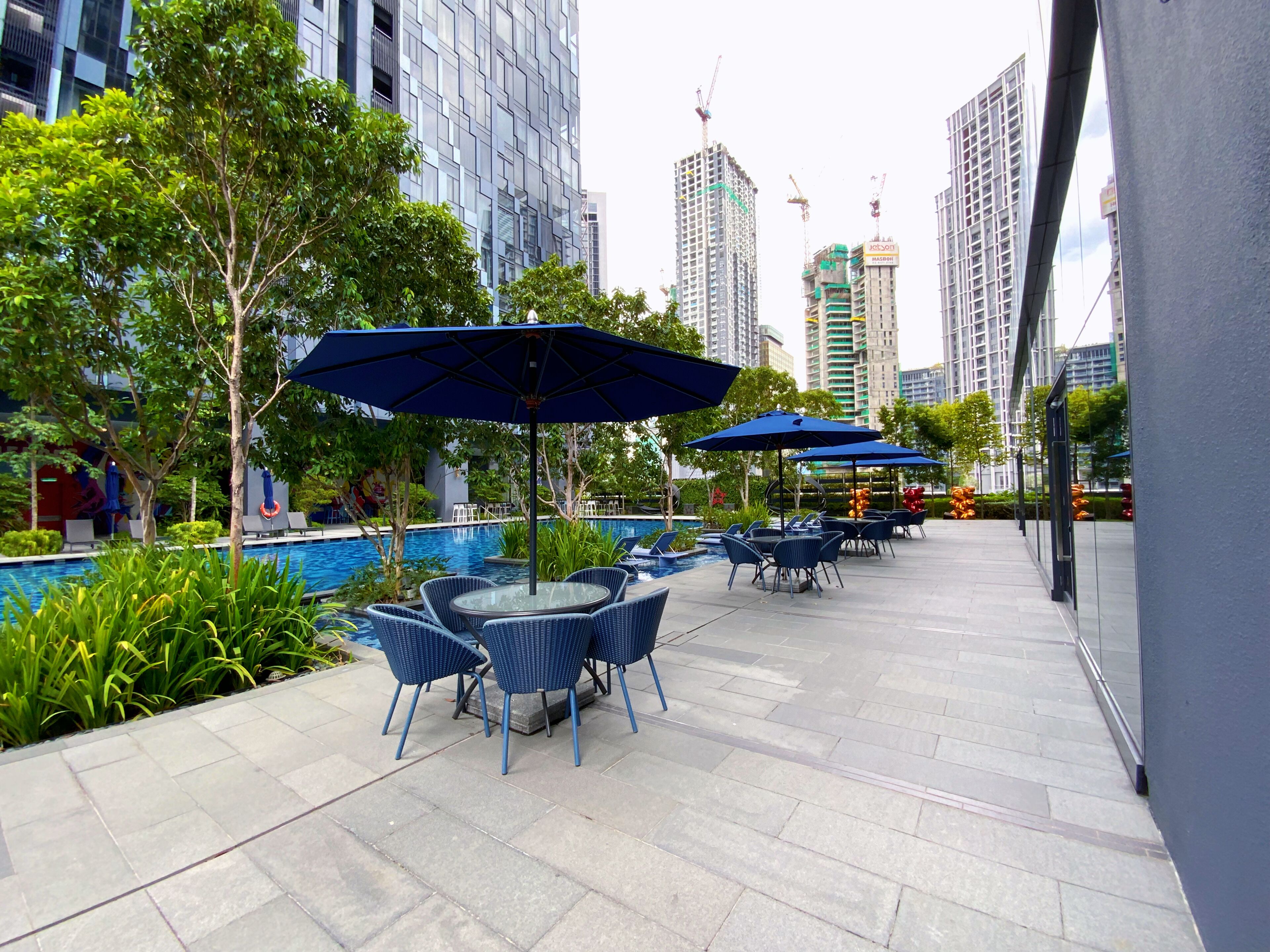 An outdoor pool area with lounge chairs and umbrellas, surrounded by greenery and modern buildings in the background.