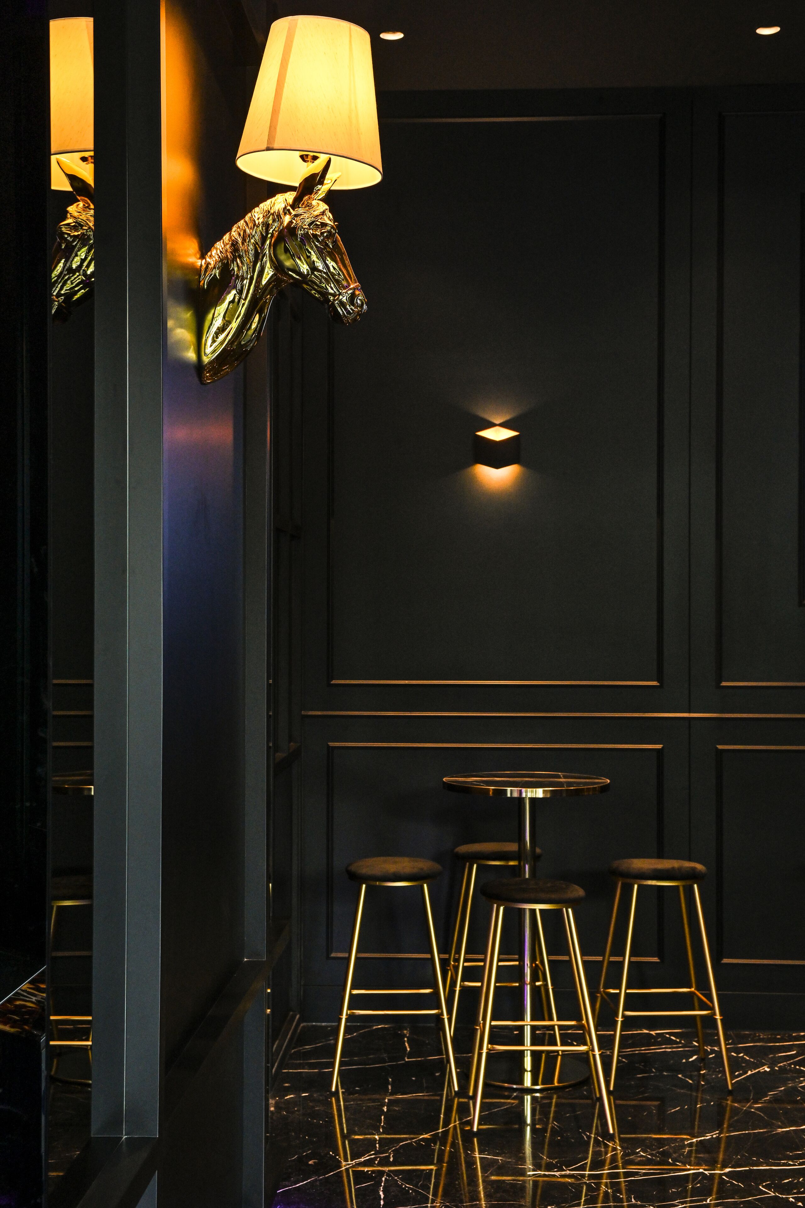 Een donker interieur met een spiegelende vloer, een lamp met gouden voet en een kruk met gouden frame.