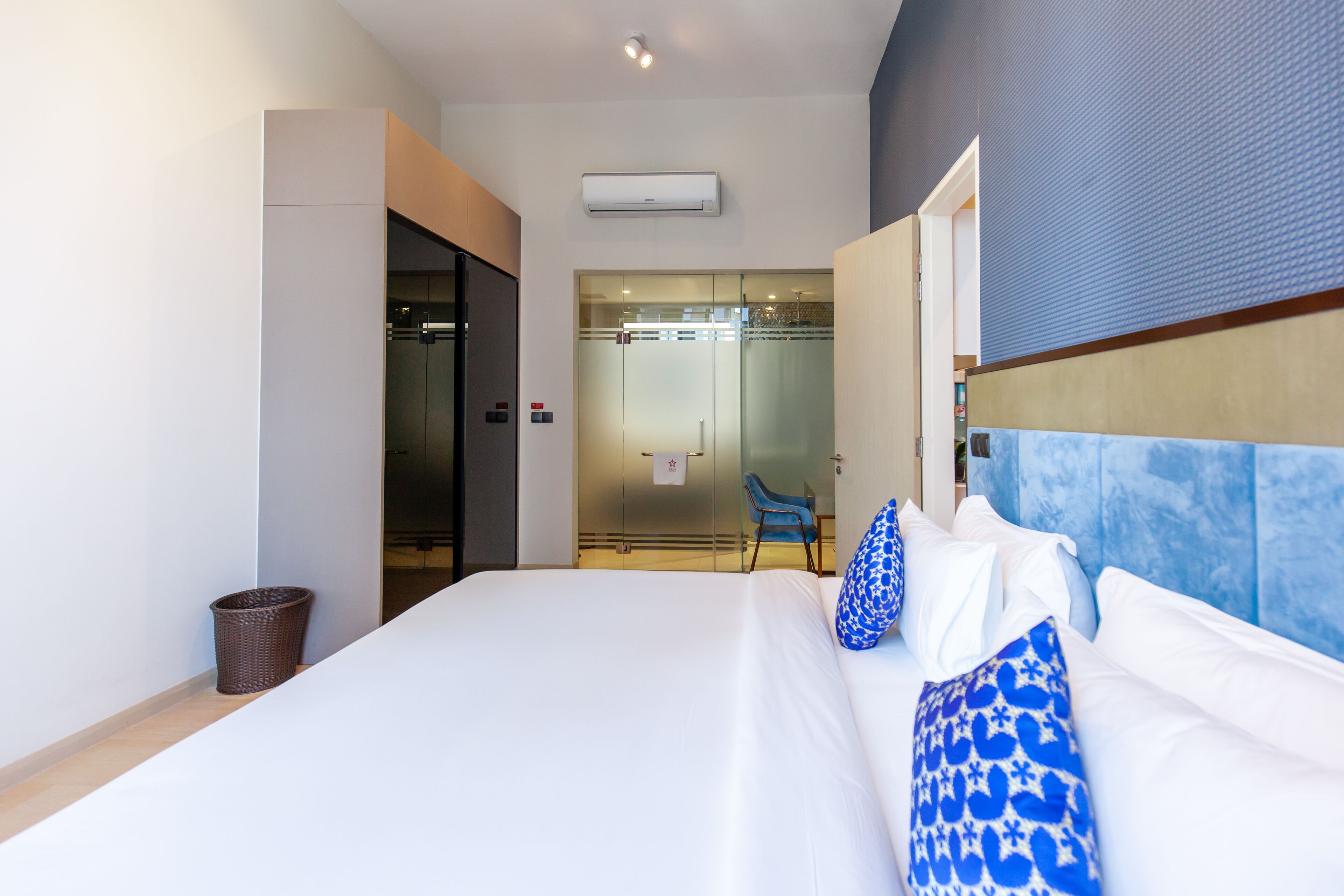 A hotel room with a large bed, a blue and white patterned headboard, a glass shower enclosure, and a small trash bin.