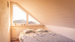 Une petite chambre confortable, avec un plafond en pente, une fenêtre donnant sur la verdure, un radiateur et un lit recouvert d’une literie à motifs floraux.