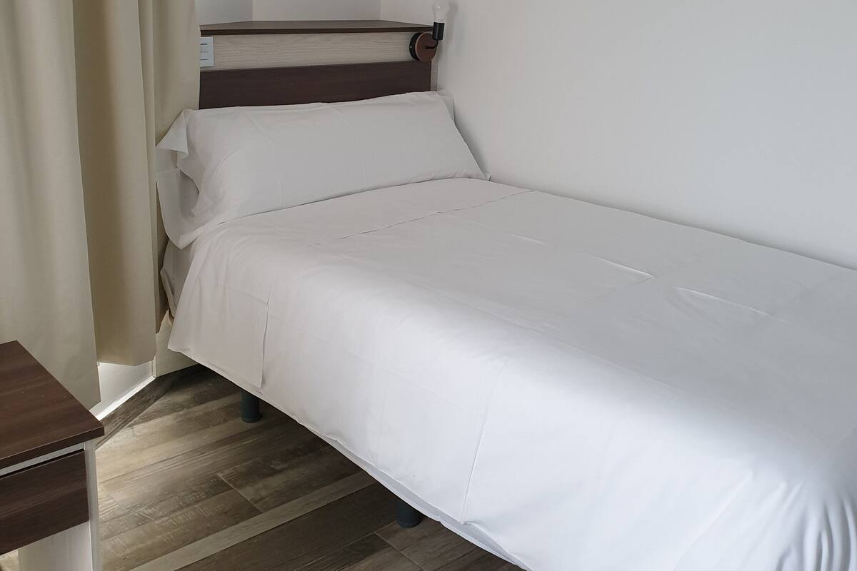 Una cama individual con ropa de cama blanca en una habitación con suelo de madera y cabecera de madera.