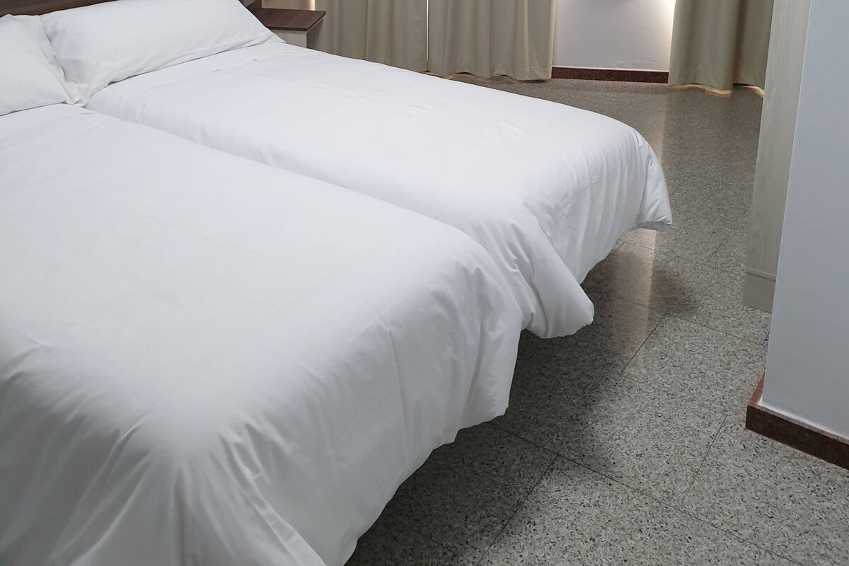 Habitación de hotel con dos camas, cabecera de madera, mesita de noche y ventana con cortinas.