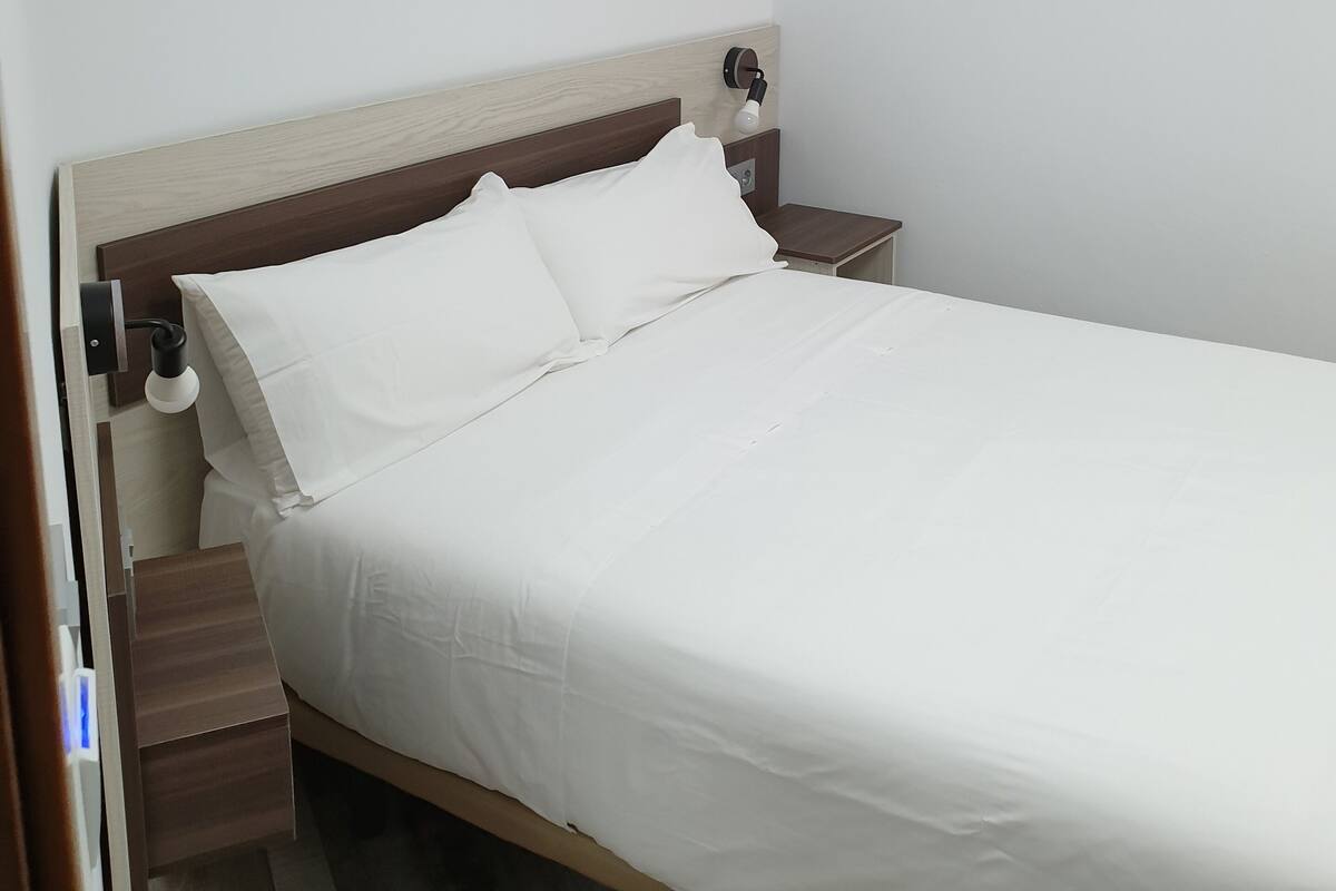 Una cama bien hecha con sábanas blancas en una habitación sencilla con cabecera y mesitas de noche de madera.
