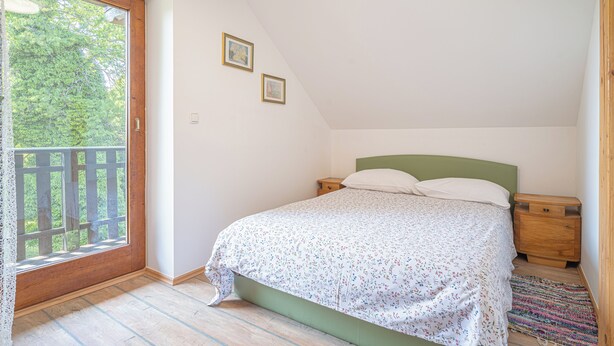 A bedroom with a bed, wooden nightstands, a green headboard, and a view through a window with a wooden railing. - Vineyard cottage Vrbekova gorca (Šmarje pri Jelšah)