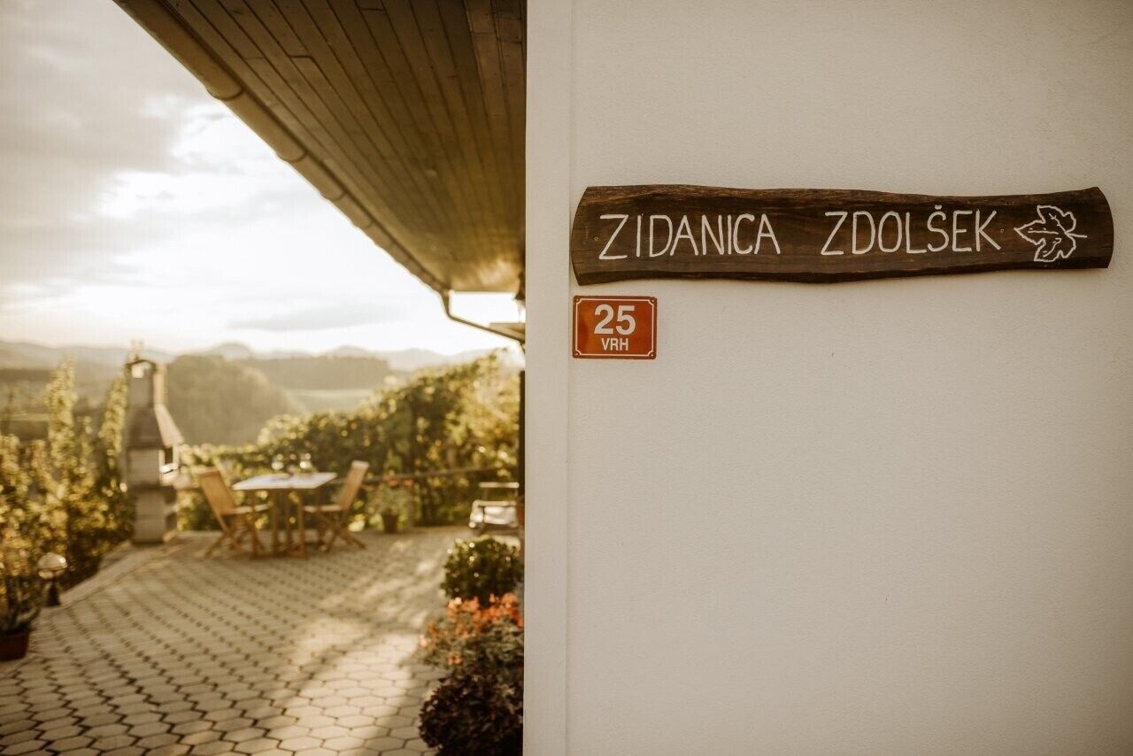 Un letrero de madera con 'Zidanica Zdolsek' y un pequeño emblema, un letrero de estacionamiento '25 VRH', y un espacio exterior pavimentado con una mesa y sillas.