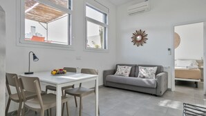 Uma sala de estar moderna com área de jantar, um sofá cinza com almofadas florais e uma cama ao fundo.