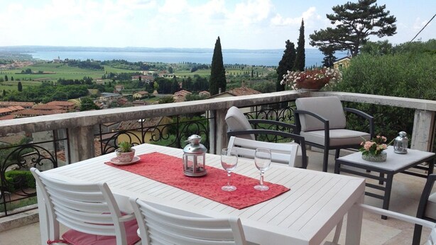 A terrace with a white table and chairs, a red placemat, and a view of a landscape with trees and a body of water. - Villa Rossa Dependance Lake Garda Bardolino-Cavaion (Dependance) (Cavaion Veronese)