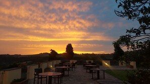 Un patio exterior con mesas y sillas, con vistas a un paisaje con árboles y un atardecer.