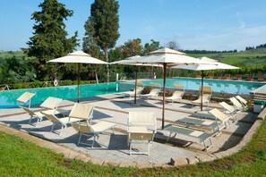 Zona junto a la piscina con sillas de descanso y sombrillas, rodeada de una piscina y vegetación.