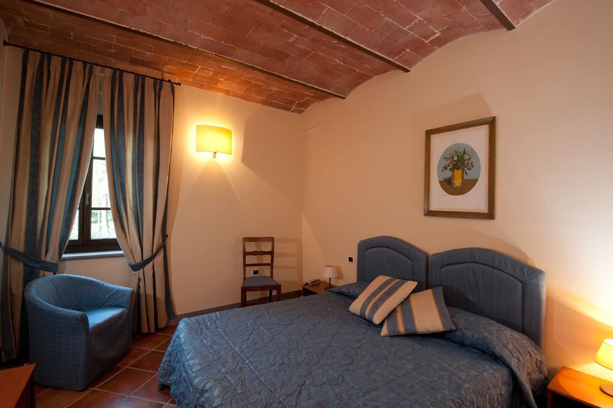 A bedroom with a bed, a chair, a nightstand, a lamp, a window with curtains, and a wall-mounted picture.