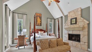 A bedroom with a four-poster bed, a stone fireplace, a large window, and a ceiling fan.