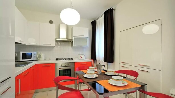 A compact kitchen with white cabinets, a gas stove, a microwave, and a dining table with four red chairs. - Red House ai Mori (Venice)