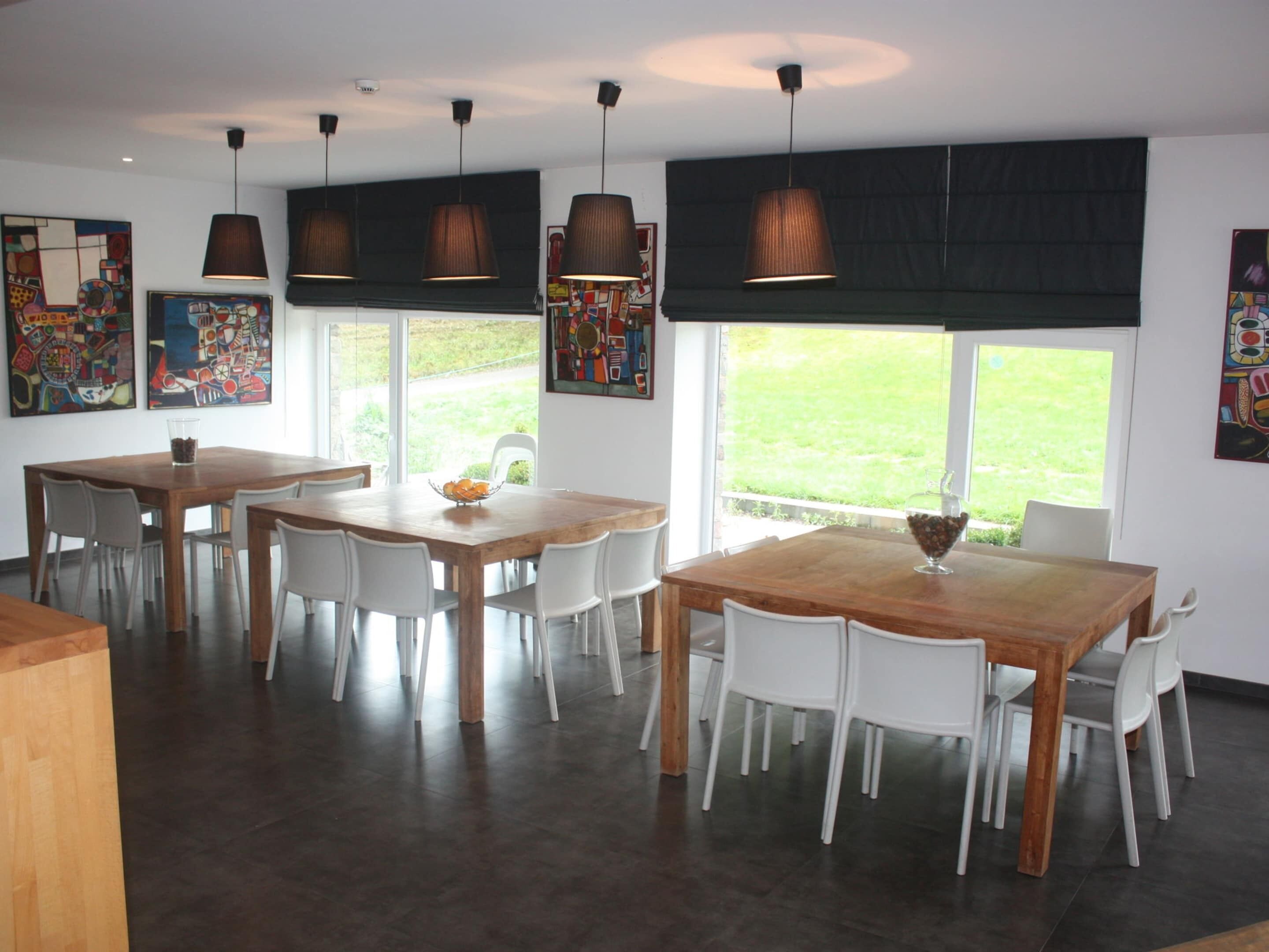 A modern dining area with wooden tables and white chairs, large windows with a view of the outdoors, and artwork on the walls.