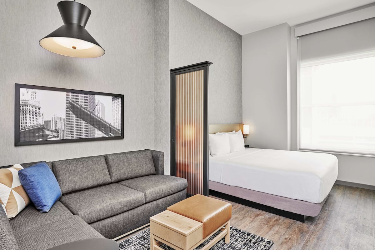 Et moderne hotelværelse med en grå sofa, en træpude, en seng med hvide sengetøj, et billed af et bybillede, en høj, lodret rumdeler og en sort pendellampe.
