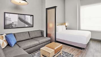 A modern hotel room with a grey sofa, a wooden ottoman, a bed with white linens, a cityscape framed picture, a tall vertical partition, and a black pendant light.