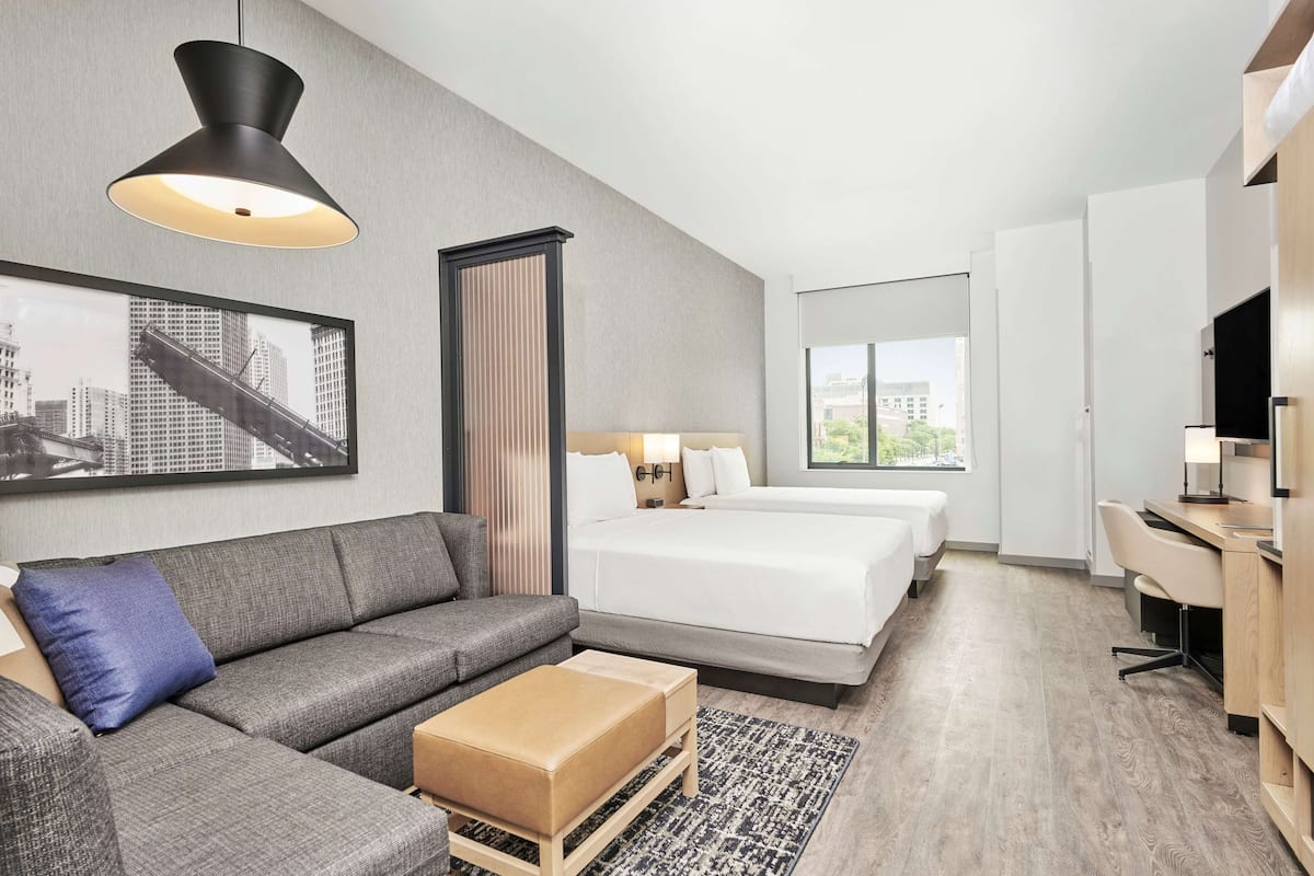 Et moderne hotelværelse med en grå sofa, seng med hvide sengetøj, et skrivebord med stol og et stort vindue med byudsigt.