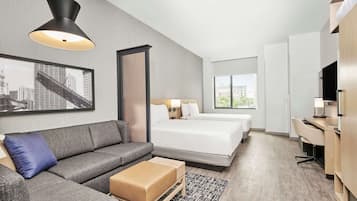 A modern hotel room with a gray sofa, a bed with white linens, a desk with a chair, and a large window offering a city view.
