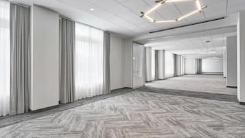 A spacious, modern hotel lobby with a herringbone-patterned carpet, large windows with grey curtains, and a geometric ceiling light fixture.