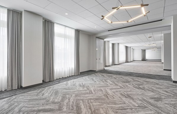 Um hall de hotel espaçoso e moderno, com carpete em padrão herringbone, grandes janelas com cortinas cinzas e um lustre de teto geométrico.