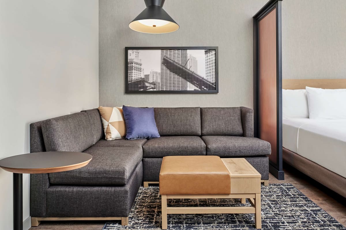 Et moderne hotelværelse med en grå modulsofa, et lille træ-sidetæppe, et rundt sidetæppe, en enkelt seng med hvide sengetæpper, et sort-hvidt bybillede på væggen og en sort hængelampe.