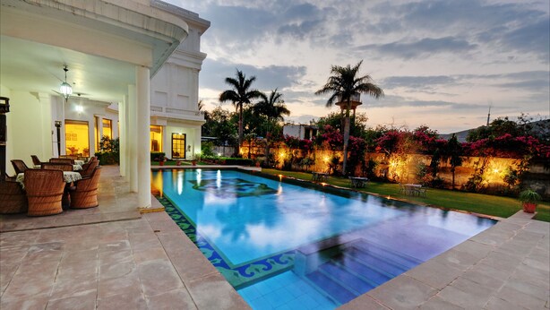 A swimming pool with a tiled deck, surrounded by a well-maintained garden with palm trees and flowering plants. - The Hadoti Palace (Bundi)