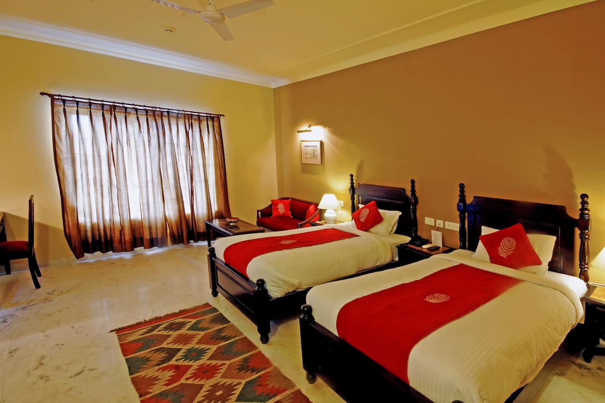 Quarto de hotel com duas camas, uma janela grande com cortinas, um ventilador de teto e uma mesa de cabeceira com abajur.
