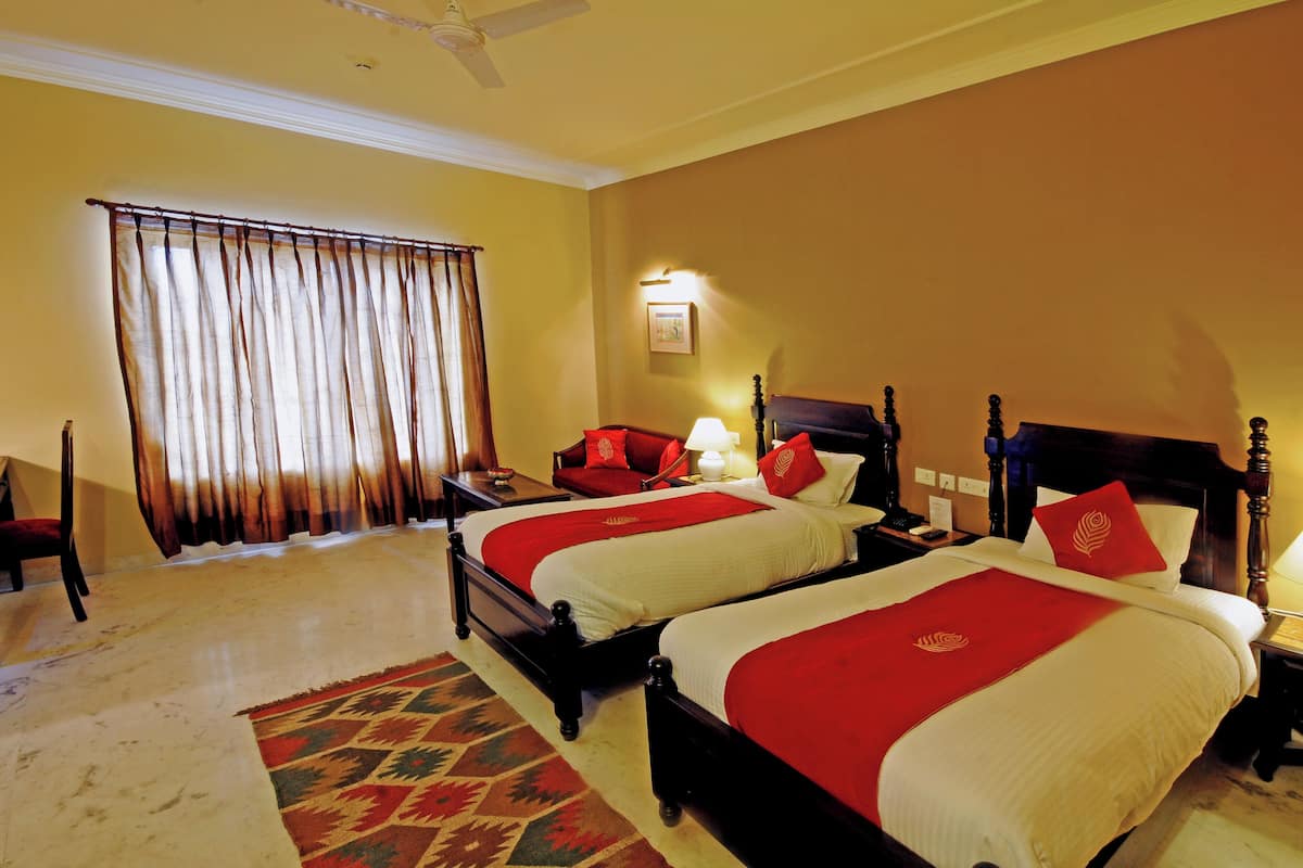 Quarto de hotel com duas camas, uma janela grande com cortinas, uma cabeceira de madeira, mesa de cabeceira com abajur e tapete.