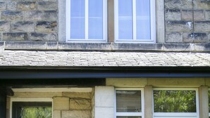 Ein Steinhaus mit einer dunklen Haustür, zwei Fenstern mit weißen Rahmen und Kübelpflanzen auf der Veranda.