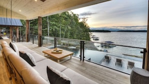 Une vaste terrasse dotée d’un plancher en bois, d’un ensemble de meubles de salon et d’une table. Une balustrade en verre offre une vue sur une étendue d’eau et des arbres.