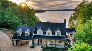 Une grande maison à deux étages, avec un toit sombre, de nombreuses fenêtres et un garage. Elle est entourée d’une végétation luxuriante et on aperçoit un lac au loin.