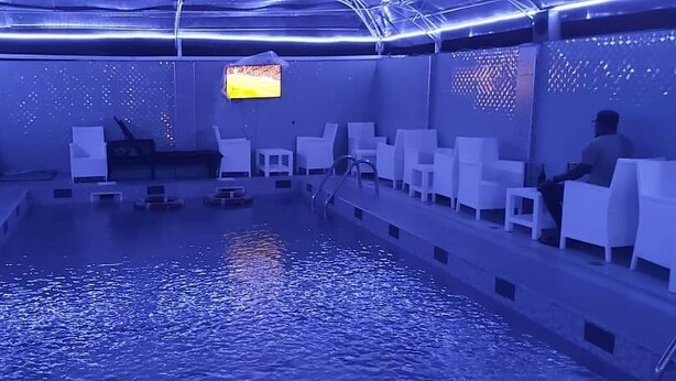 A pool area with white chairs arranged around the perimeter, a television mounted on the wall, and a ceiling with blue lighting. - Seafortunes Royal Suites (Lagos)