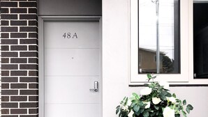 Entrada moderna de un edificio con una puerta blanca marcada '48A', un ventanal con vistas y dos plantas en macetas.