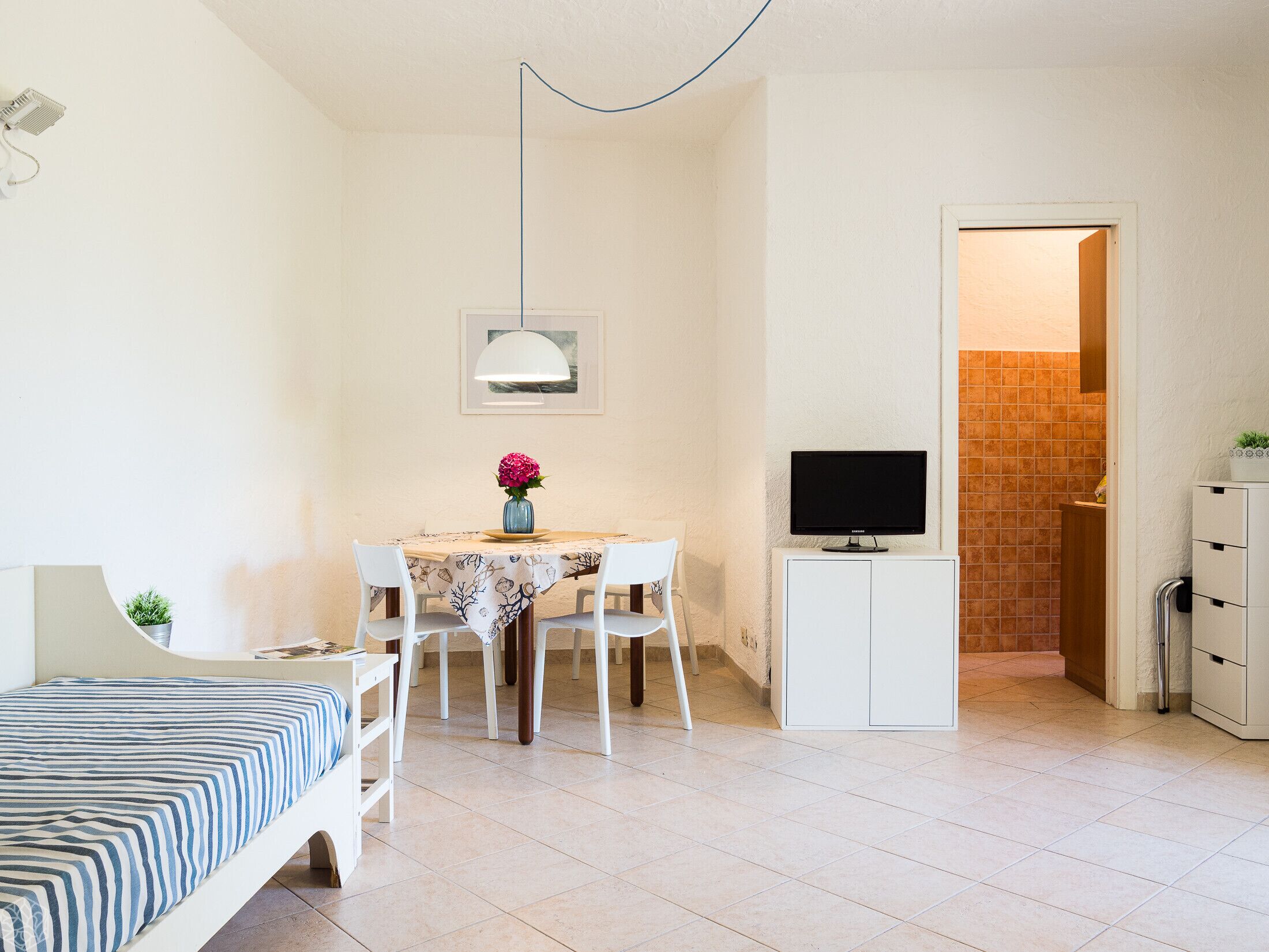 A compact bedroom with a bed, a small table with a vase of flowers, a TV on a cabinet, and a bathroom visible through an open door.