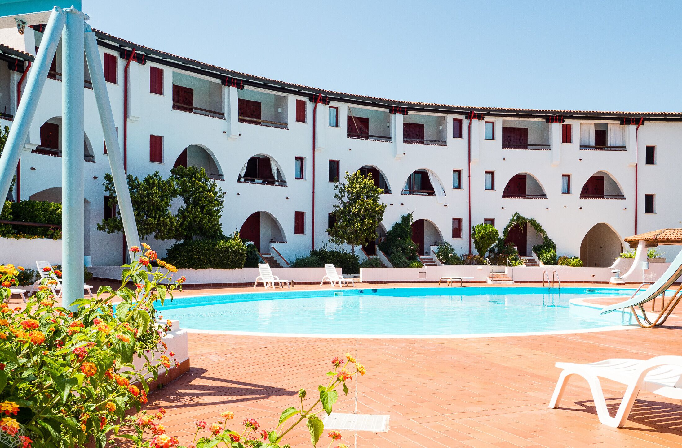 A pool area with a white building, red-framed windows, and a clear blue sky.
